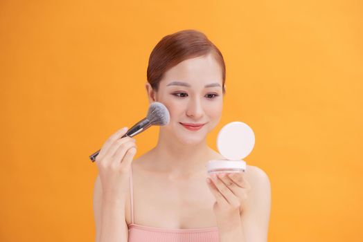 Beautiful woman applying blusher to her cheek with a large cosmetics brush while holding a compact mirror in front of her