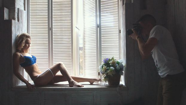 Young blonde model in the blue underwear posing for photographer sitting at the window during fashion photo session, shooting in the studio