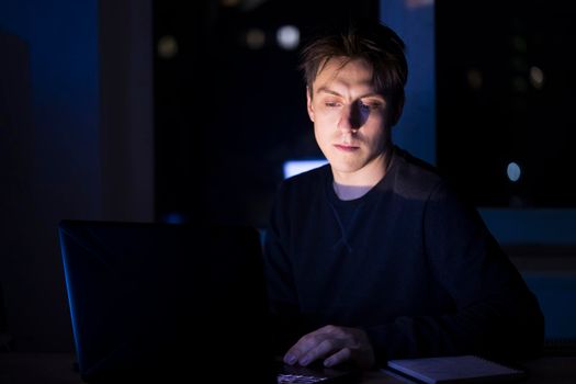 A young man sits and works at night on a laptop on the network, types on the keyboard, writes text messages, studies, prepares a project, a programmer, web designer works as a freelancer at home.