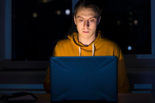 A young man sits and works at night on a laptop on the network, types on the keyboard, writes text messages, studies, prepares a project, a programmer, web designer works as a freelancer at home.