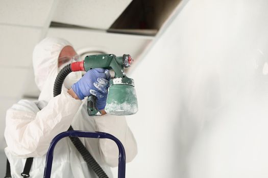 Close-up of male worker painting wall with spray gun in white colour, busy handyman at work. Renovation, construction site, interior, household concept