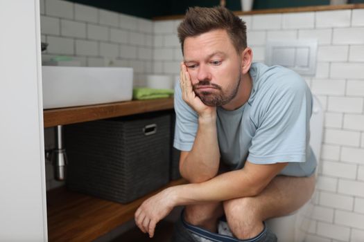 Portrait of bored man on toilet seat dream about something, fulfill natural need, male in bathroom with modern interior. Wc, hygiene, nature calls concept