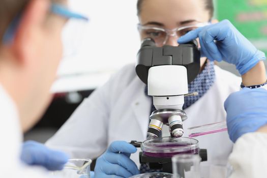 Close-up of female scientist explore purple sample under microscope in laboratory. Male chemist add liquid from flask. Lab, science, investigation concept