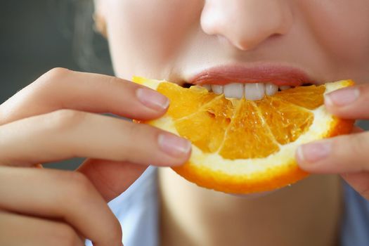 Close-up of woman hold in hand slice of chopped orange eats for breakfast with mouth in kitchen. Healthy eating, diet, juicy, fruit, summer harvest concept