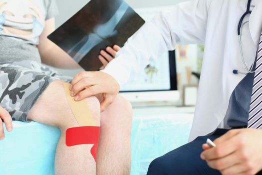 Close-up of physician wear uniform and stethoscope carefully examining patient leg. Client come with scan of bone. Kinesiology tape on knee. Injury concept