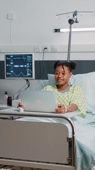 Young patient using video call on laptop for remote communication while sitting in hospital ward bed. Woman talking to friends on online video conference conference about healthcare.