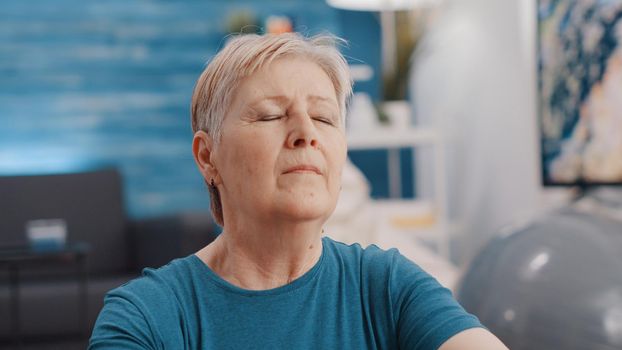 Close up of old woman feeling zen with eyes closed at home. Portrait of senior person relaxing after meditation on yoga mat. Retired adult meditating for wellness and calm recreation