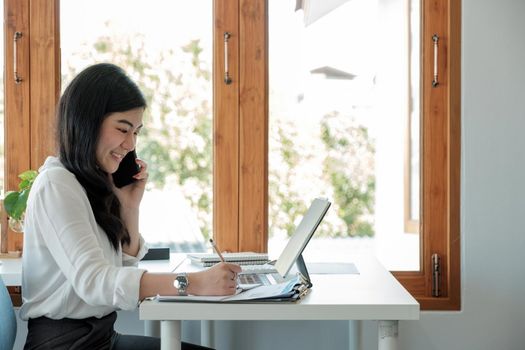 Side view happy asian businesswoman sitting on her workplace in the office. Young woman working for financial document data charts with laptop during call phone in the office.