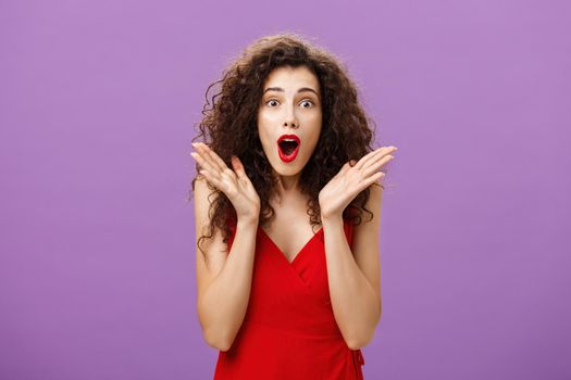 Waist-up shot of excited and amazed expressive elegant adult. curly-haired woman with red lipstick in evening luxurious dress gasping from amazement waving palms near face staring surprised at camera.