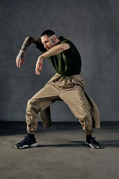 Young stylish performer with tattooed body and face, earrings, beard. Dressed in khaki t-shirt and overalls, black sneakers. Dancing on gray background. Dancehall, hip-hop. Full length, copy space