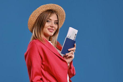 Cute blonde model in straw hat, white blouse and red pantsuit. She is smiling, holding passport and ticket, posing sideways against blue studio background. Travelling concept. Close-up, copy space