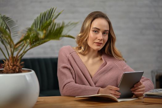 Blonde lady in pink cardigan is holding a tablet. Sitting at wooden table with notebooks and palm tree in pot on it. Student, blogger. Interior with white brick wall and gray sofa. Close-up