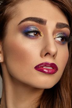 Brunette woman with luxury makeup and perfect skin is looking aside. Multi-colored eyeshadow, false eyelashes, glossy burgundy lips. Professional maquillage. Close up