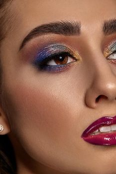 Brunette female with luxury makeup and perfect skin is looking away. Blue and golden eyeshadow, long eyelashes, glossy burgundy lips and white teeth. Professional maquillage. Close up