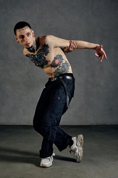 Athletic guy with tattooed body and face, naked torso, beard. Dressed in black pants and white sneakers. He is dancing against gray studio background. Dancehall, hip-hop. Full length, copy space