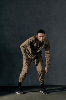 Young stylish male with tattooed body and face, earrings, beard. Dressed in khaki jumpsuit and black sneakers. He is dancing against gray studio background. Dancehall, hip-hop. Full length, copy space