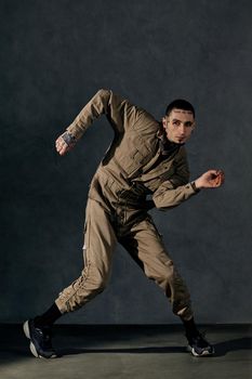 Young stylish fellow with tattooed body and face, earrings, beard. Dressed in khaki jumpsuit and black sneakers. He dancing against gray studio background. Dancehall, hip-hop. Full length, copy space