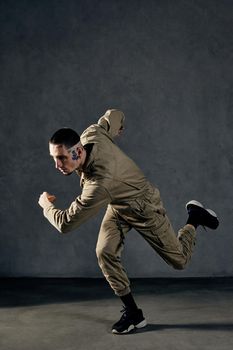 Young stately fellow with tattooed body and face, earrings, beard. Dressed in khaki jumpsuit and black sneakers. He dancing against gray studio background. Dancehall, hip-hop. Full length, copy space