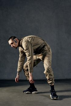 Young strong man with tattooed body and face, earrings, beard. Dressed in khaki overalls and black sneakers. He is dancing against gray studio background. Dancehall, hip-hop. Full length, copy space