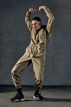 Young active guy with tattooed body and face, earrings, beard. Dressed in khaki overalls and black sneakers. He is dancing against gray studio background. Dancehall, hip-hop. Full length, copy space