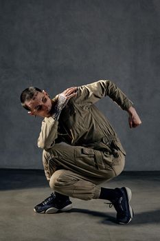 Extraordinary man with tattooed body and face, earrings, beard. Dressed in khaki overalls and black sneakers. He is dancing against gray studio background. Dancehall, hip-hop. Full length, copy space