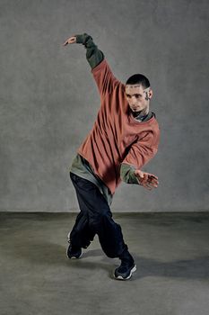 Stately fellow with tattooed body and face, earrings, beard. Dressed in colorful jumper, black pants and sneakers. Dancing on gray background. Dancehall, hip-hop. Full length, copy space