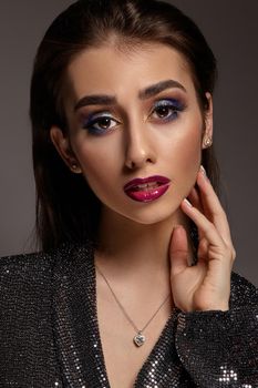 Brunette model in black shiny dress and jewelry is touching her face, posing on gray background. Luxury makeup, perfect skin. Multi-colored eyeshadow, long eyelashes, glossy red lips. Close up