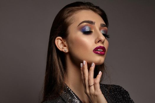 Brunette woman in jewelry and black shiny dress is touching her face, posing on gray background. Luxury makeup. Colorful eyeshadow, false eyelashes, glossy red lips. Professional maquillage. Close up