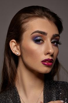 Brunette female in jewelry and black shiny dress is looking at you, posing on gray background. Luxury makeup. Colorful eyeshadow, false eyelashes, glossy red lips. Professional maquillage. Close up