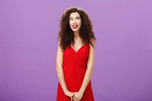 Studio shot of cute thoughtful and creative charming european woman in stylish red licking lips with desire and amusement looking at upper right corner smiling, daydreaming using imagination. Copy space