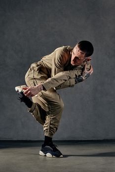 Young attractive man with tattooed body and face, earrings, beard. Dressed in khaki overalls and black sneakers. He dancing against gray studio background. Dancehall, hip-hop. Full length, copy space