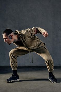 Young unusual man with tattooed body and face, earrings, beard. Dressed in khaki overalls and black sneakers. He is dancing against gray studio background. Dancehall, hip-hop. Full length, copy space