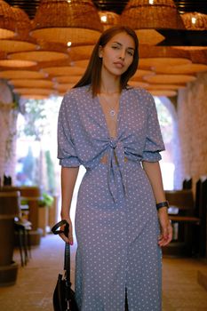 Adorable blonde maiden in a long blue dress with polka-dots, watch, with a pendant around her neck and a small black handbag in her hand is looking at the camera, posing standing under a huge chandeliers in a city cafe while walking alone. The concept of fashion and style. Close-up shot.