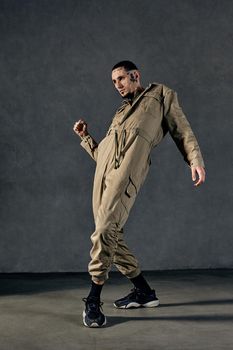 Young flexible fellow with tattooed body and face, earrings, beard. Dressed in khaki jumpsuit and black sneakers. He dancing against gray studio background. Dancehall, hip-hop. Full length, copy space
