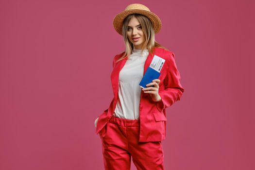 Gorgeous blonde lady in straw hat, white blouse and red pantsuit. She put her hand in pocket, smiling, holding passport and ticket, posing on pink background. Travelling concept. Close-up, copy space