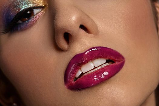 Face of a lady with luxury makeup and perfect skin. She is looking at you. Blue and golden eyeshadow, long eyelashes, glossy burgundy lips and white teeth. Professional maquillage. Close up