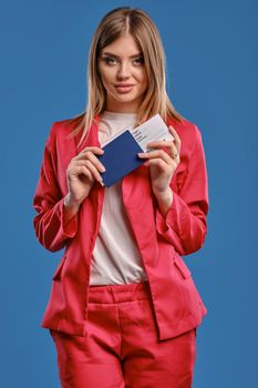 Alluring blonde woman in white blouse and red pantsuit. She is smiling, holding passport and ticket, posing against blue studio background. Travelling concept. Close-up, copy space