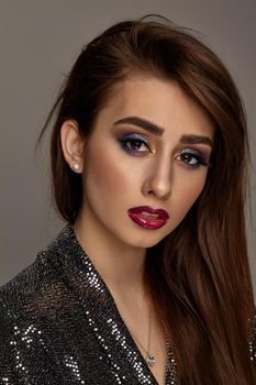 Brunette model in black shiny dress and jewelry posing against gray background. Luxury makeup, perfect skin. Colorful eyeshadow, false eyelashes, glossy red lips. Professional maquillage. Close up