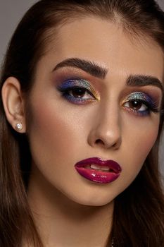 Brunette girl in earrings looking at you while posing on gray background. Luxury makeup, perfect skin. Multi-colored eyeshadow, false eyelashes, glossy purple lips. Professional maquillage. Close up