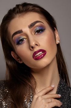 Brunette female in black shiny dress and jewelry has opened her mouth, posing on gray background. Luxury makeup, perfect skin. Multi-colored eyeshadow, false eyelashes, glossy pink lips. Close up