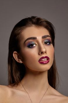 Brunette naked woman in necklace and earrings is posing against gray studio background. Luxury makeup, perfect skin. Colorful eyeshadow, long eyelashes, glossy purple lips and white teeth. Close up