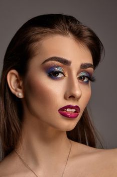 Brunette naked female in necklace and earrings is posing against gray studio background. Bright makeup, perfect skin. Colorful eyeshadow, long eyelashes, glossy red lips and white teeth. Close up