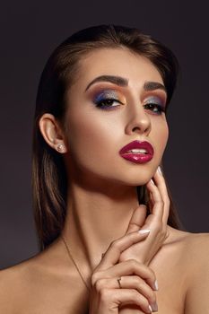 Brunette naked lady in jewelry is touching her face, posing against gray studio background. Luxury makeup, perfect skin. Colorful eyeshadow, false eyelashes, glossy red lips and white teeth. Close up