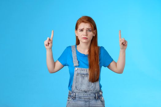 Doubtful concerned unsure redhead female making important decision, grimacing hesitant displeased, point up promo, top copy space, feel uncertain unlikely buy suspicious product, blue background.