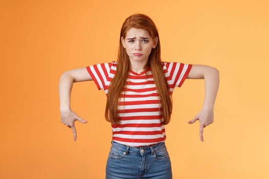Cute timid hesitant ginger female taking tough decision feel pressured upset, frowning, pulling gloomy unhappy face, pointing down disappointed, uneasy taking decision, orange background.