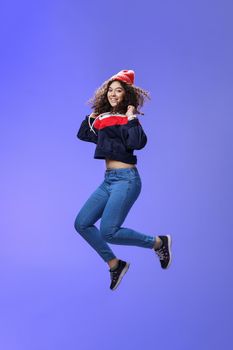 Studio shot of cute girl with curly hair in beanie sweatshirt and sneakers jumping playful and carefree over blue background, having fun enjoying cool weather smiling broadly as flying in air.