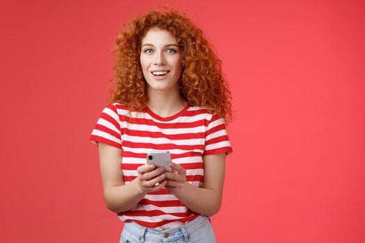 Fashionable carefree good-looking relaxed redhead girlfriend hold smartphone look camera excited pleased turn attention friend answering messages social media writing post red background.