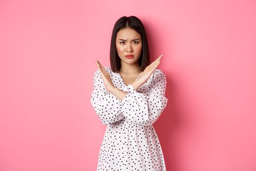 Angry asian woman tell stop, making cross to prohibit or disapprove something, frowning displeased, standing over pink background.