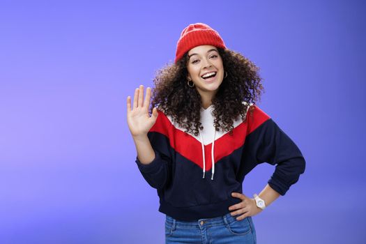 Hey nice to meet you. Charming female ski couch in cute red beanie with curly hair waving hello with palm and smiling broadly greeting newbies as teaching winter sports over blue background. Copy space