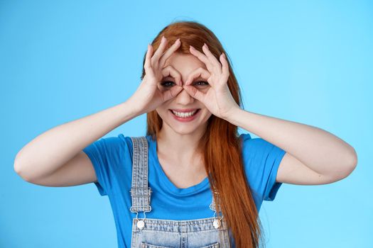 Playful cheerful redhead european woman, long nature red hairstyle, show okay finger glasses look through ok signs, smiling broadly funny grimace, stand blue background carefree enjoy summer.